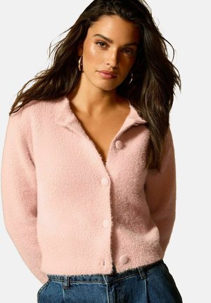 Cardigan rosa fuzzy con colletto, chiusura a bottoni frontale e maniche lunghe. Tessuto testurizzato con una vestibilità rilassata e lunghezza cropped, abbinato a jeans.