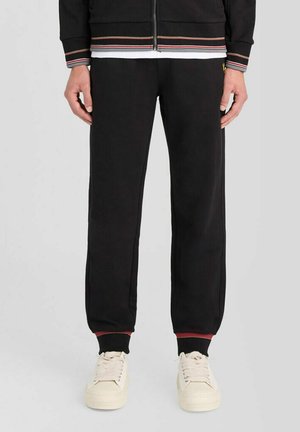 Person iført sorte joggingbukser med ribbet kant, en sort lynlåsjakke og off-white sneakers, stående på en ensfarvet baggrund.