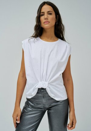 Top blanco de manga corta con un frente anudado, que presenta pequeños adornos dispersos. Combinado con pantalones ajustados plateados brillantes.