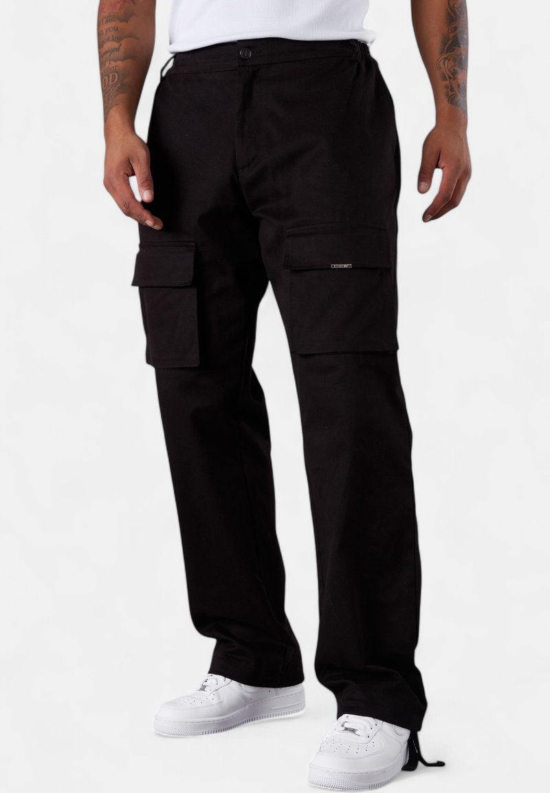 Schwarze Cargo-Hosen aus robustem Stoff, mit mehreren Seitentaschen, entspannter Passform und verstellbaren Kordelzügen an den Bündchen, kombiniert mit weißen Sneakers.