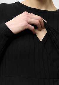 Μαύρο ριντζέ ύφασμα με μακριά μανίκια, με άνοιγμα σε σχήμα V. Ένα χέρι τραβάει το ντεκολτέ, αποκαλύπτοντας ένα εσωτερικό στρώμα.