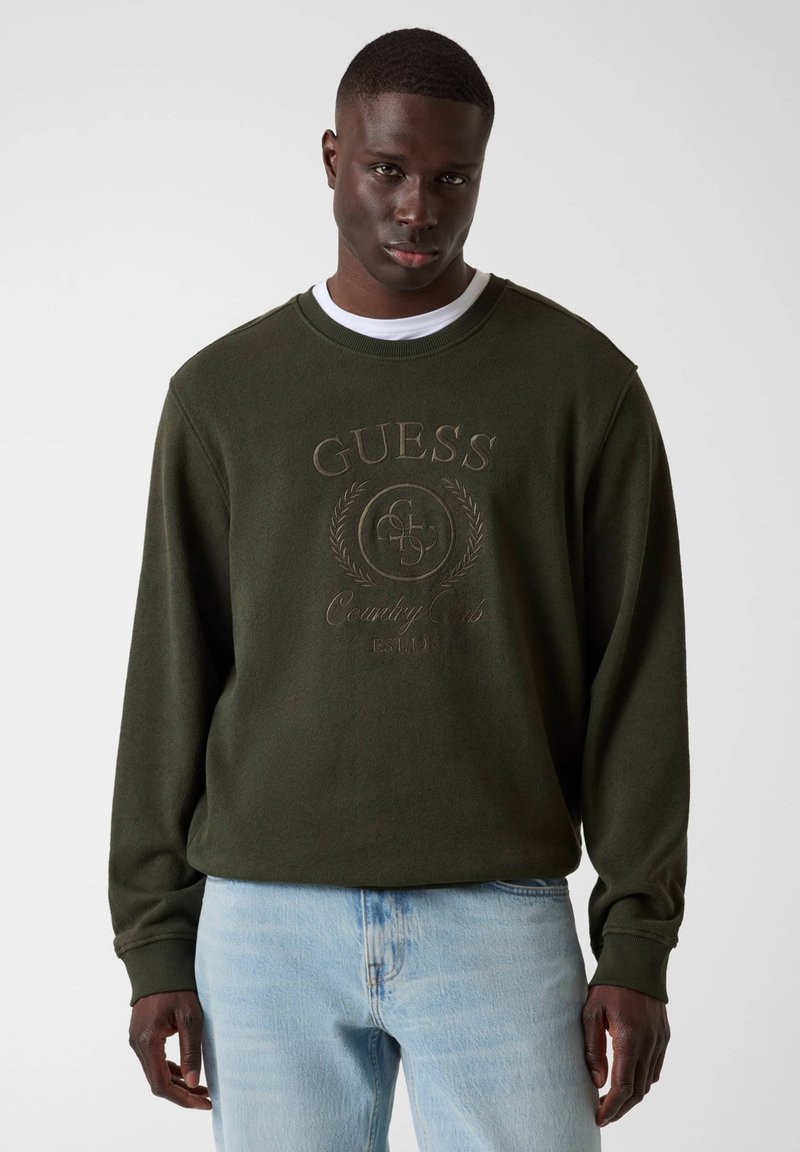 Homme portant un sweat-shirt Guess vert foncé avec logo embossé et jean bleu clair, debout devant un fond gris uni.