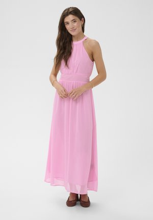 Mladá žena s dlhými hnedými vlasmi, oblečená v ružových maxi šatách bez rukávov so zaväzovaním za krkom, obutá v hnedých pletených balerínach, stojaca so zloženými rukami.