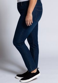 Tmavě modré džíny s úzkým střihem, s elastickým pasem a standardními zadními kapsami, spárované s černými nazouvacími botami.