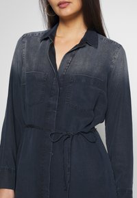 Abito chemisier in denim blu scuro con chiusura a bottoni, due tasche sul petto, maniche lunghe e dettaglio in vita per stringere.