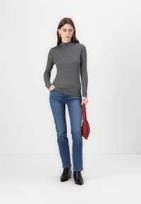 Top gri cu mâneci lungi și guler înalt, asociat cu blugi drepți de culoare albastră deschisă. Modelul ține o geantă roșie și poartă cizme cu toc negre.