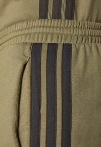 Olivenfargede joggebukser med ribbestrikket liv, sidelommer og tre svarte vertikale striper langs ytre søm. Mykt, teksturert stoff.