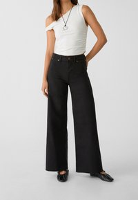 Zwarte wijde jeans met hoge taille, gecombineerd met een wit top met één schouder en zwarte platte schoenen; minimale hardware details zichtbaar.