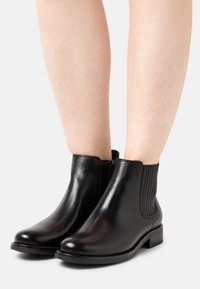 Černé kožené kotníkové boty s hladkým povrchem, bočními elastickými panely a nízkým masivním podpatkem, s kulatou špičkou a minimálními kovovými doplňky.