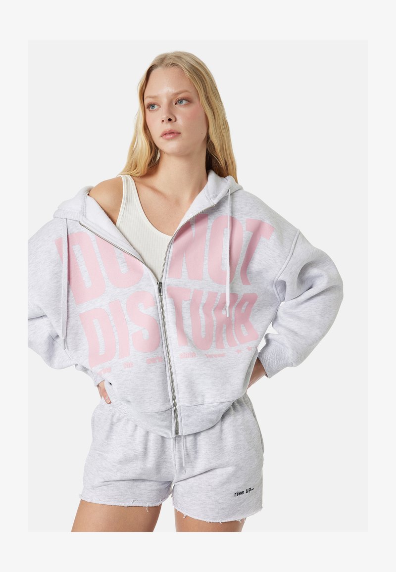 Joven mujer con sudadera con capucha gris claro con cremallera con texto rosa "NO MOLESTAR" y pantalones cortos a juego con texto "rise UP" en la pierna.