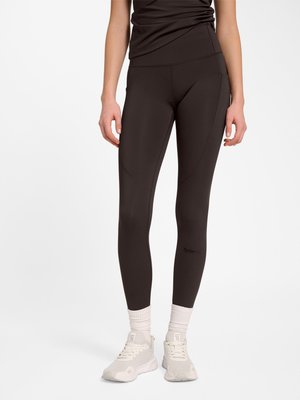 Kvinde iført sorte tætsiddende leggings, hvide sokker og hvide sportssko, stående mod en ensfarvet hvid baggrund.
