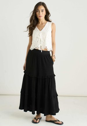 Woman in sleeveless white eyelet top and long black tiered skirt wearing black flip-flops, standing against plain light background.