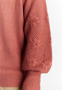 Maglione lavorato a maglia in rosa polveroso, caratterizzato da sezioni testurizzate e lisce. Manica a sbuffo decorata con accenti floreali ricamati. Polsini a costine.