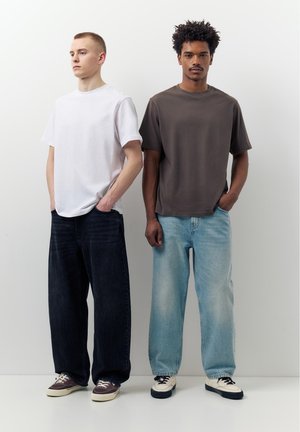 Két férfi áll egymás mellett egy fehér fal előtt, az egyik fehér pólót és sötét farmert visel, a másik barna pólót világoskék farmerrel.