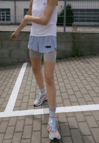 Osoba ve bílém tílku Adidas, modrých vzorovaných šortkách, béžových cyklistických šortkách, pruhovaných ponožkách a běžeckých botách stojí na asfaltované ploše se bílými čarami.