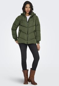 Puffer jacket in dark green, with a zippered front, hood, and elastic cuffs. Worn with black skinny jeans and brown leather knee-high boots.