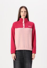 Fleece pullover in roze en rood, met een hoge kraag, knopen aan de voorkant en een GAP-logo. Zachte textuur met een kleurblokontwerp.