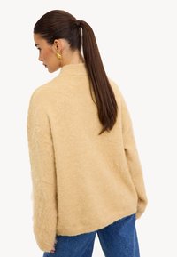 Pull en beige duveteux avec un col montant et une coupe ample, doté de manches texturées et d'un ourlet sans coutures, associé à un jean bleu.