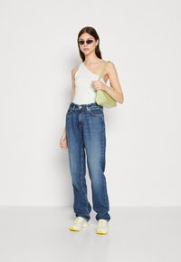 Top blanco de un solo hombro, vaqueros de tiro alto azules, zapatillas con acentos amarillos y un bolso pequeño verde. La modelo lleva gafas de sol negras.