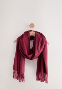 Tiefroter Schal mit Fransenenden, der auf einem hellen Holzbügel vor einer schlichten weißen Wand drapiert hängt.