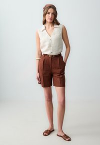 Camicia senza maniche in lino beige con bottoni, pantaloni corti in lino marrone con una cintura e sandali alla schiava marroni. Capelli raccolti con un foulard a motivo marrone.