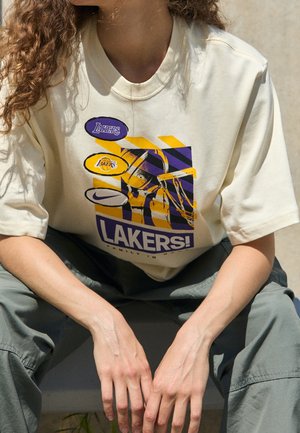 Osoba z kręconymi włosami w kremowej koszulce Lakers z fioletowymi i żółtymi wzorami, siedząca z rękami opartymi między szarymi spodniami.