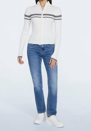 Femme portant un pull côtelé blanc zippé avec des rayures grises, un jean bleu à jambes droites et des baskets blanches, debout devant un fond uni.