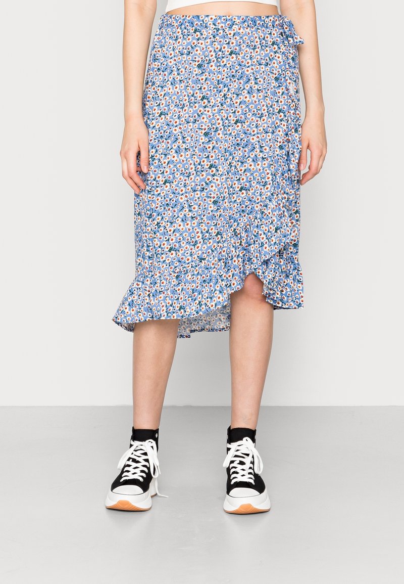 Femme portant une jupe midi fleurie bleue avec un ourlet à volants et des baskets montantes noires et blanches, debout sur un fond uni.