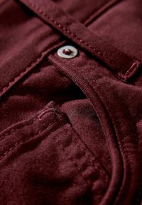 Bordeaux stof met knoop- en stikseldetails, zichtbaar zaknaad en zachte textuur. Subtiele glans verbetert het uiterlijk.