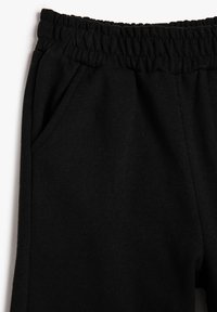 Pantalones de cintura elástica negra con detalle de bolsillo lateral sobre un fondo blanco, mostrando la sección superior izquierda de la prenda.