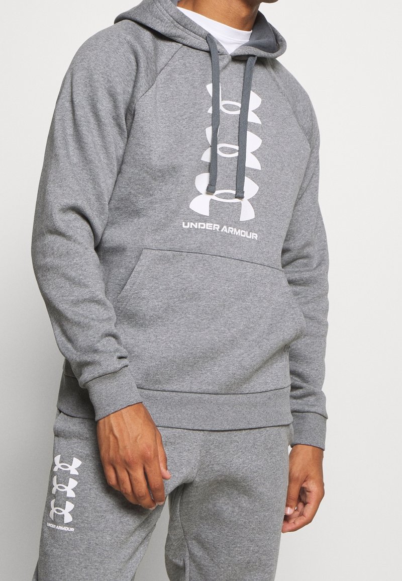 Homme portant un sweat à capuche gris Under Armour et un pantalon de survêtement assorti avec un logo blanc et du texte, debout devant un fond clair uni.