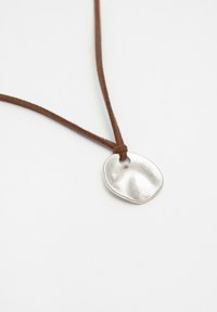 Pendente in argento con una forma organica e texture, appeso a un cordino in suede marrone, legato a metà.