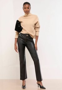 Sieviete ģērbusies bēša krāsas augstās apkakles džemperī, melnās ādas biksēs, melnās augstpapēžu kurpēs un tur melnu somiņu zem rokas.