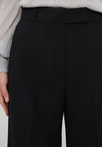 Mustriga mustad püksid siledast kangast, lameda eesotsaga ja peente voldikutega, koos halli pika varrukaga topiga.