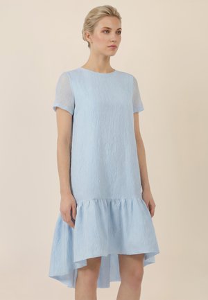 Blondynka stoi, ubrana w teksturowaną, jasnoniebieską sukienkę z krótkim rękawem i falbanowym, asymetrycznym dołem na beżowym tle.