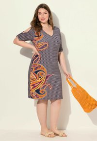 Vestido de manga corta en color navy con un estampado de paisley en naranja y púrpura; diseño de escote en V. La modelo sostiene un bolso tejido naranja. Sandalias con detalle de nudo.