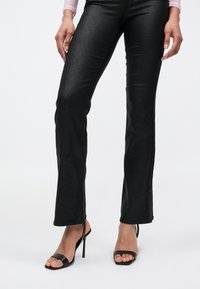 Pantalon noir évasé et brillant avec une texture lisse ; porté avec des sandales à talons hauts noires ouvertes. Le design présente une silhouette ajustée.