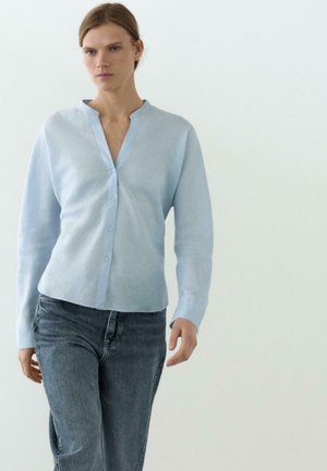 Kobieta w jasnoniebieskiej koszuli zapinanej na guziki i ciemnoszarych dżinsach idzie na tle jednolitego białego tła.