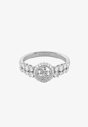 Srebrn prsten Guess s okruglim logom u sredini okružen malim prozirnim kamenjem, dodatni kamenčići duž stranica prstena.