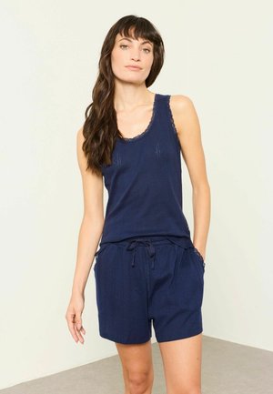 Kvinde med langt brunt hår iført en navyblå ærmeløs top og matchende shorts, der står mod en lys baggrund.