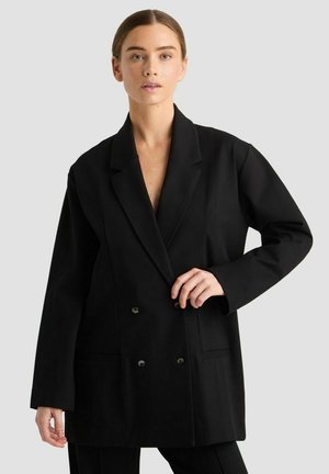 Frau mit braunen Haaren, die einen schwarzen, übergroßen, doppelt geknöpften Blazer trägt und den Blazerausschnitt mit der rechten Hand gegen einen schlichten grauen Hintergrund hält.