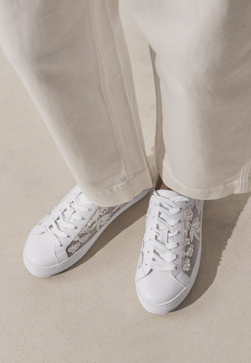 Zapatillas blancas con cordones y detalles florales de malla, combinadas con pantalones beige claro sobre un fondo neutro de suelo.
