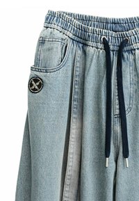 Pantalones de mezclilla azul claro con cinturilla elástica, cordón marino, bolsillos laterales y un parche de logo circular negro en el bolsillo izquierdo.