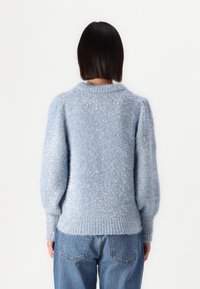 Femme avec des cheveux noirs raides portant un pull en peluche bleu clair et un jean, vue de dos sur fond blanc uni.