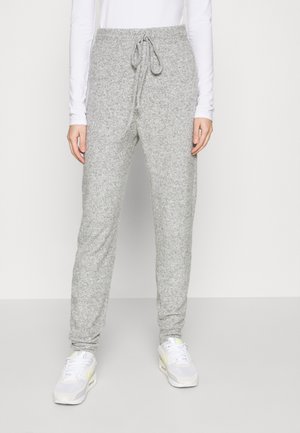 Persona que lleva pantalones jogger de punto gris claro con cintura de cordón y zapatillas blancas, de pie contra un fondo liso y claro.