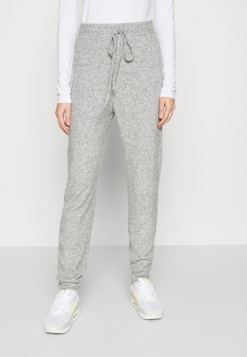Personne portant un pantalon de jogging en tricot gris clair avec une taille à cordon et des baskets blanches, debout devant un fond clair uni.