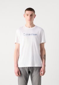 Baltas kokvilnas T-krekls ar apaļu kakla izgriezumu un īsām piedurknēm. Priekšā rakstīts zilā krāsā "Calvin Klein." Nēsāts ar pelēkiem džinsiem.