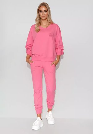 Różowa bluza z kapturem i pasujące dresy z miękkiego materiału, z ribbrowanymi mankietami i wyszywanym detalem na klatce piersiowej. Białe sneakersy dopełniają całość.