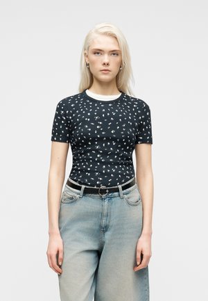 Junge Frau mit langen blonden Haaren, die ein schwarzes, kurzärmeliges Oberteil mit Blumenmuster, hellblaue, hoch taillierte Jeans und einen schwarzen Gürtel mit runder Schnalle trägt.