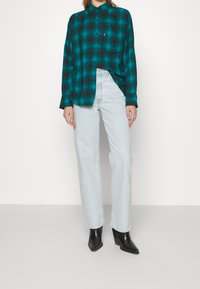 Camicia a maniche lunghe a quadri turchese e nera, con chiusura a bottoni, indossata con jeans dritti azzurri e stivaletti neri con tacco.